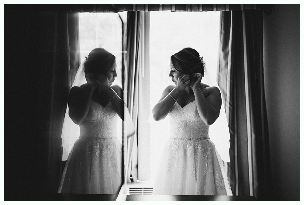 Bride putting on earrings in front of a window, reflected in a mirror. Black and white.