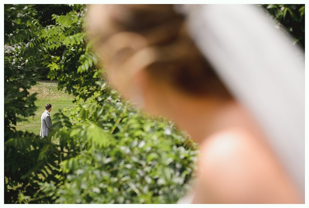 Bride in foreground, groom in the distance, waiting for first look in a sunny garden.