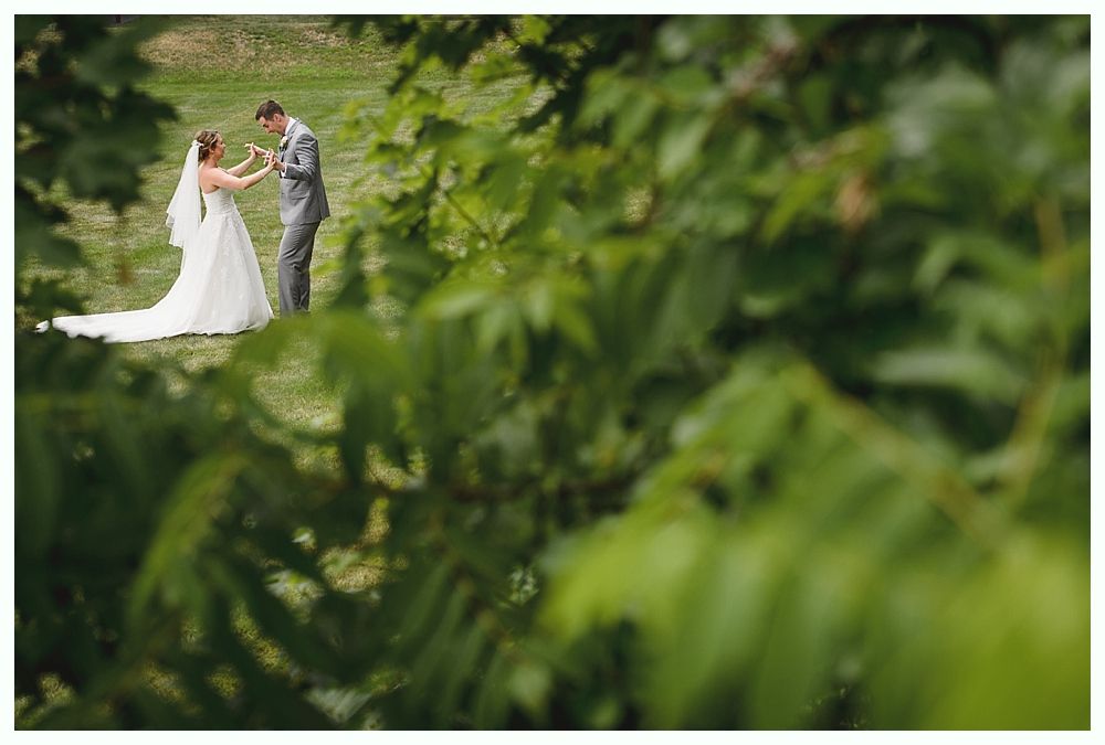 Bride and groom holding hands, dancing in a grassy field. Green foliage in foreground, soft lighting.