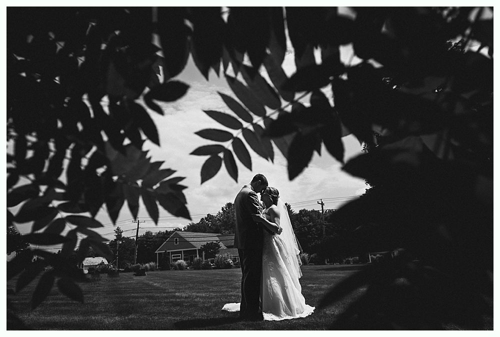 Couple embraces outdoors, framed by leaves. Groom in suit, bride in veil, near a building.