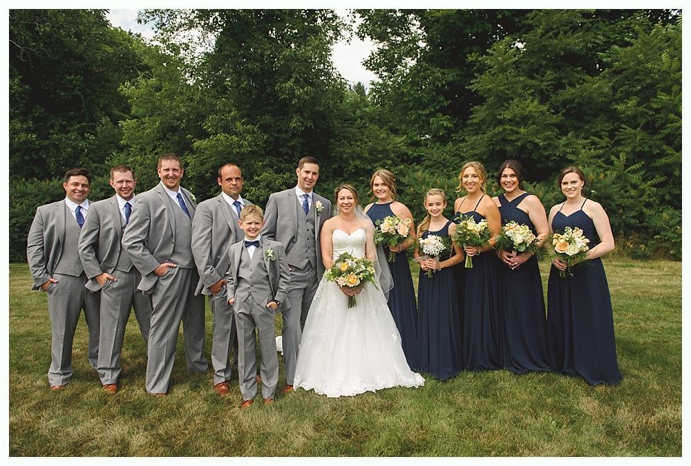 Wedding party poses outdoors. Bride in white gown, bridesmaids in navy. Groomsmen in gray suits.