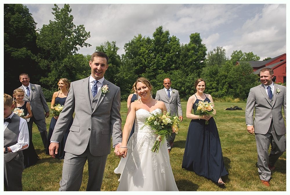 Wedding party walking in a garden. Bride and groom holding hands, smiling. Bridesmaids in navy dresses, groomsmen in gray suits.