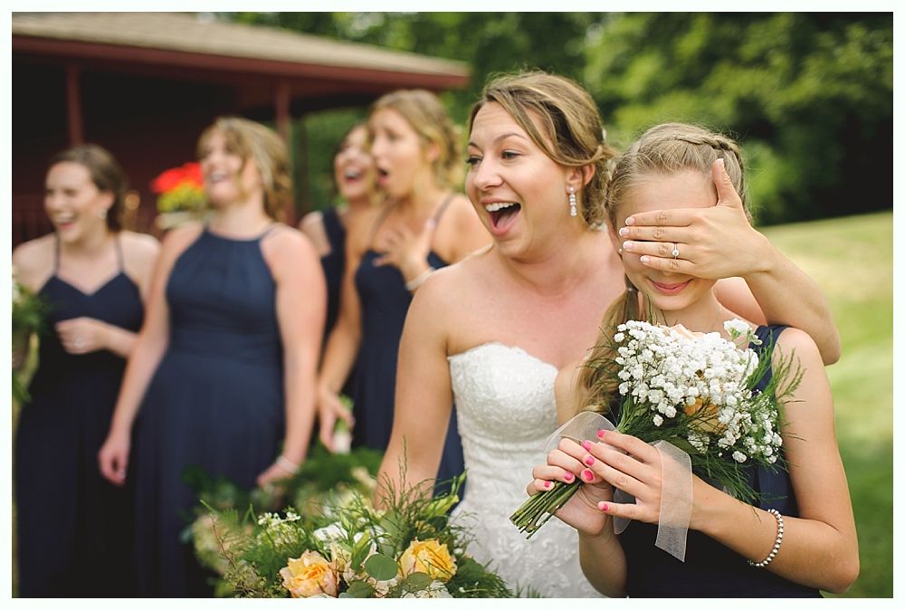 Bride surprises flower girl; bridesmaids watch, laughing; outdoor setting.