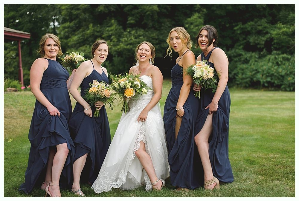 Bride and bridesmaids in navy dresses playfully lift skirts, laughing in a grassy setting.