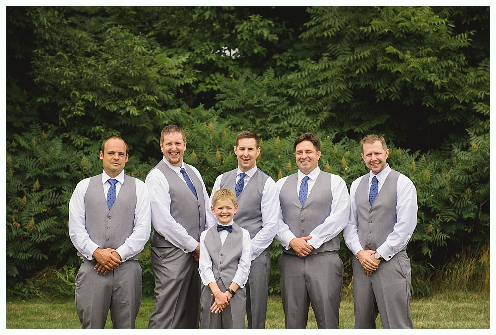 Groomsmen in gray vests and blue ties pose in front of green foliage. One younger boy wears a bow tie.
