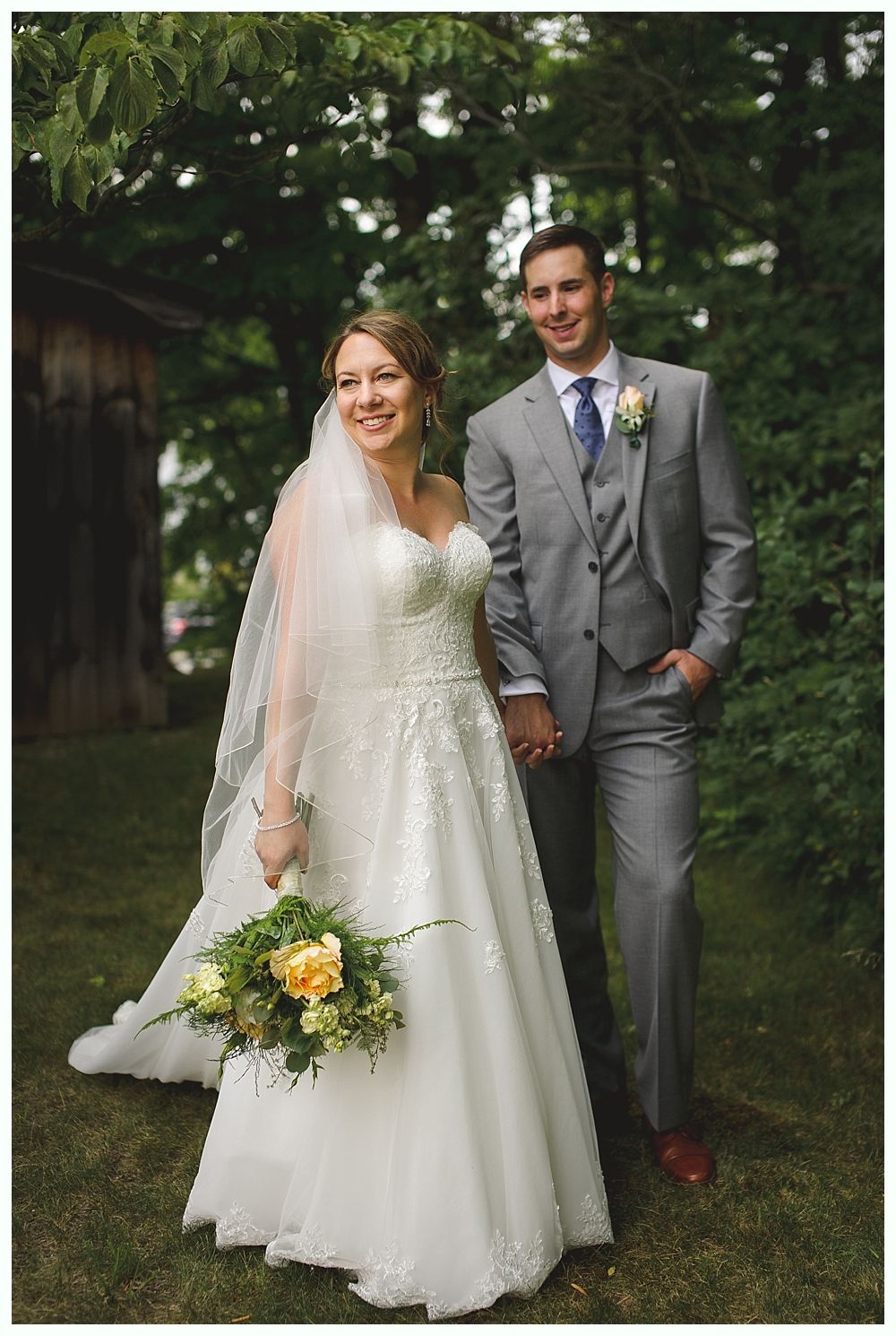 Bride and groom smiling, holding hands. Bride in white gown with veil, groom in gray suit, outdoors.