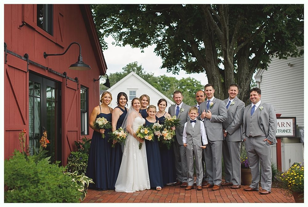 Wedding party poses in front of a red barn. Bride in white gown with bridesmaids in blue dresses, groomsmen in gray suits.