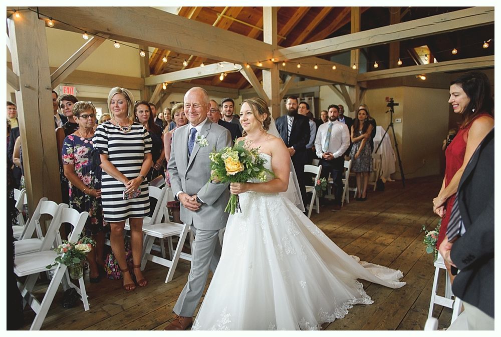 Bride walks down aisle with father at a wedding ceremony in a rustic venue. Guests watch.
