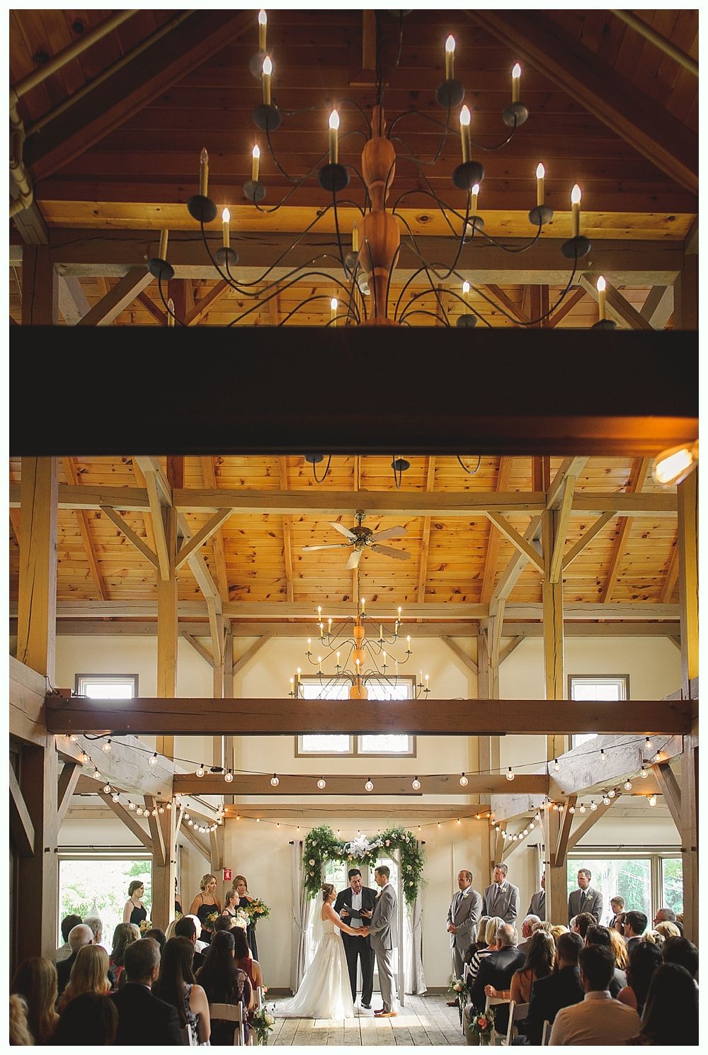 Wedding ceremony in a rustic barn with couple, guests, floral arch, and chandelier.