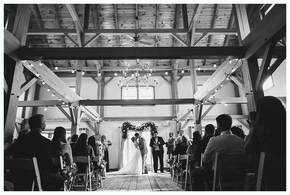 Wedding ceremony in a barn; couple kissing under an archway, surrounded by guests.