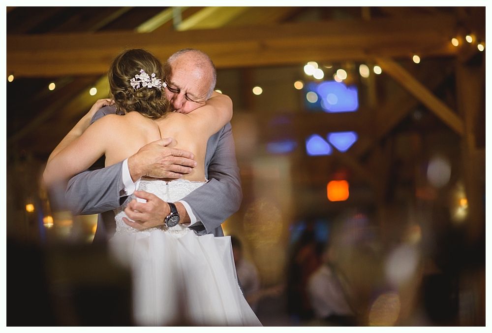 Bride and father dance, embracing at a wedding reception. Barn setting with string lights and blurred guests.