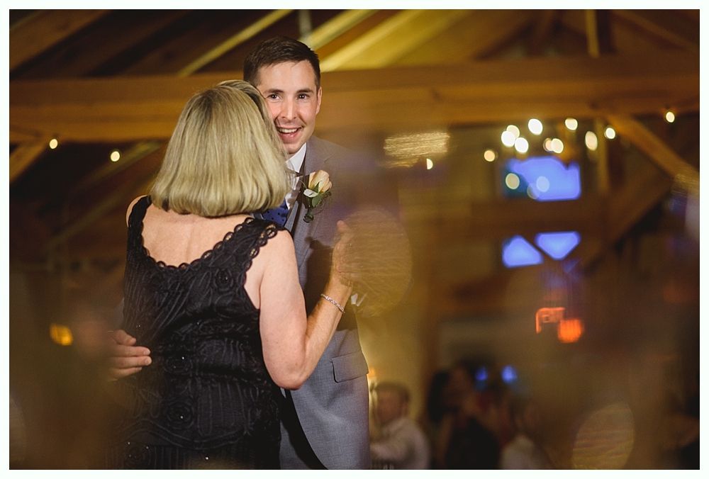 Man dances with a woman in a black dress at a wedding reception. They smile; lights and decorations in background.