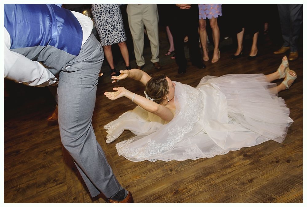 Bride on the floor in wedding dress, arms outstretched, next to a person in a vest and slacks on a wood floor.