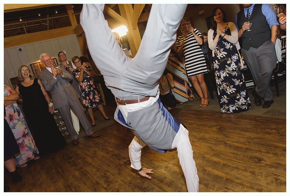 Man in formal wear doing a handstand on a wooden dance floor, guests watching.