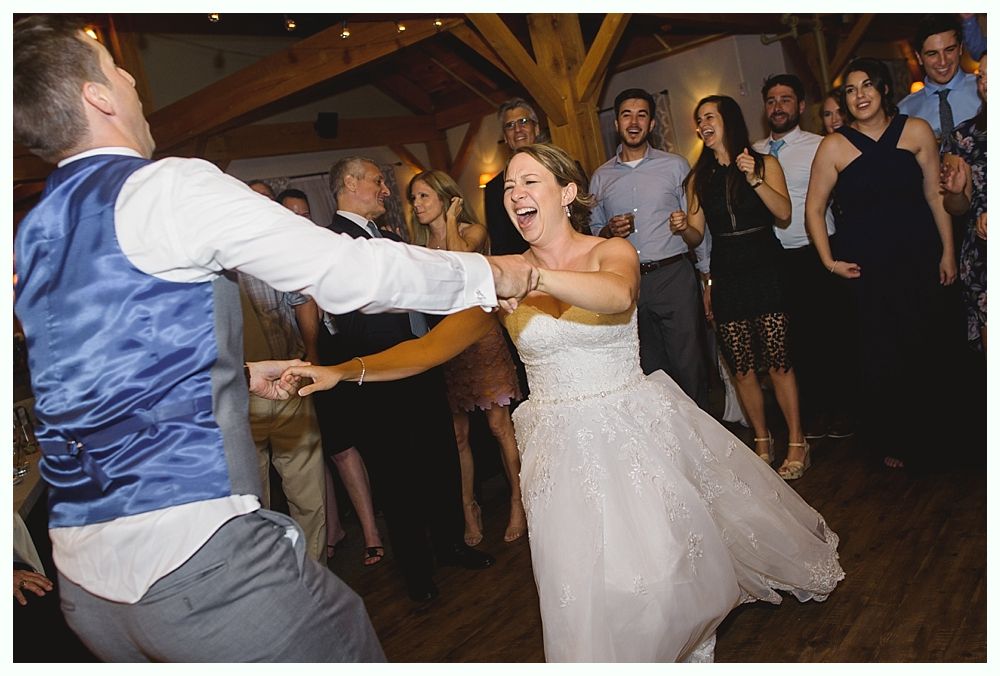 Bride and groom dancing at a wedding reception; she is laughing, wearing a white dress, and he wears a blue vest.