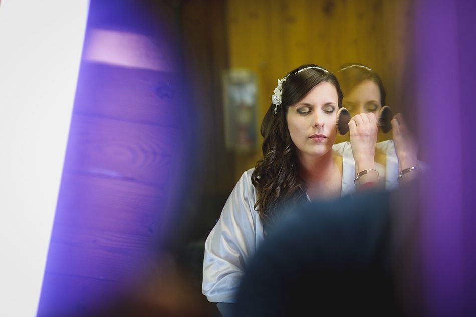 Bride applying makeup in front of a mirror, wearing a white robe and a floral headpiece; soft purple and white hues.