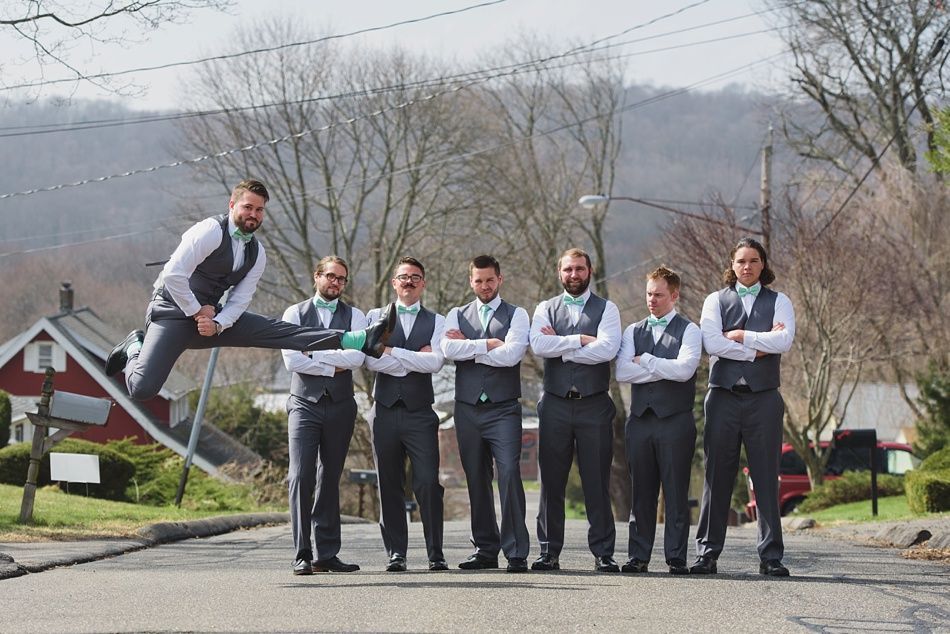 Groomsmen pose on a street; one man jumps in the air. All wear gray vests and bow ties, arms crossed.