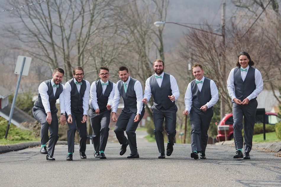 Men in gray vests and pants, teal bow ties, and socks, pose playfully on a street with a mountainous backdrop.