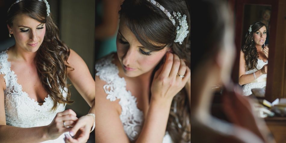Bride in a white lace dress putting on earrings, looking in a mirror with curly brown hair.