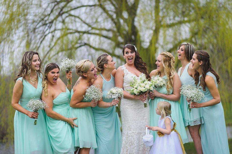 Bride and bridesmaids pose outdoors, all in turquoise dresses, reacting with surprise; flower girl nearby.