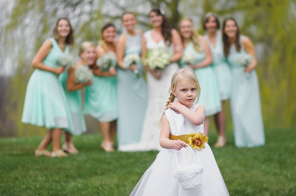 Flower girl in white dress with gold sash, looks at camera. Bridesmaids in mint green dresses and bride in white in background.