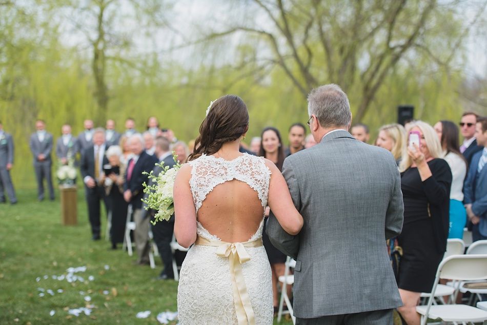 Bride walking down aisle with father, backless dress, outdoor ceremony. Guests and wedding party present.