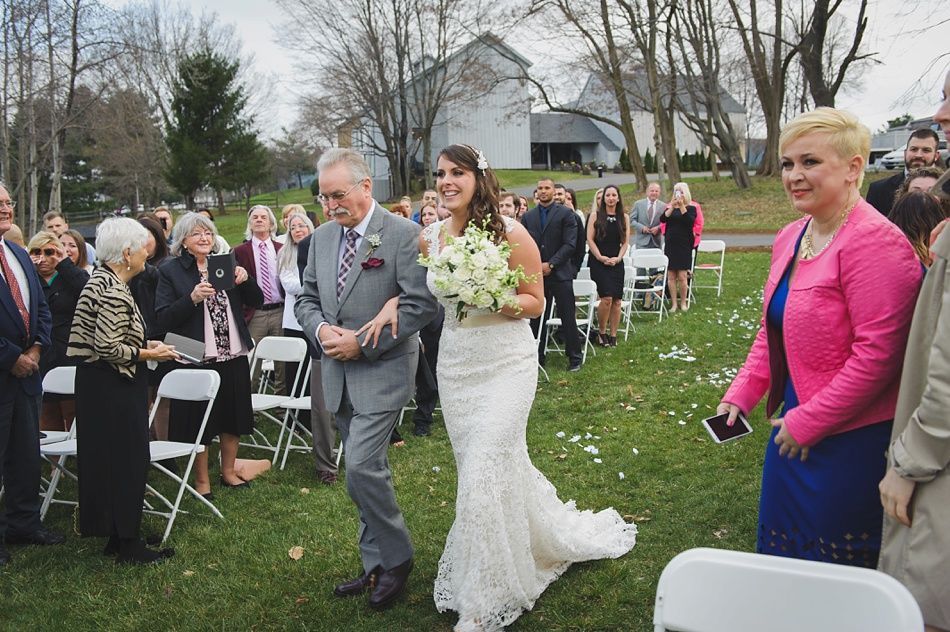 Bride walking down aisle with father, holding bouquet, at outdoor wedding. Guests seated on lawn.