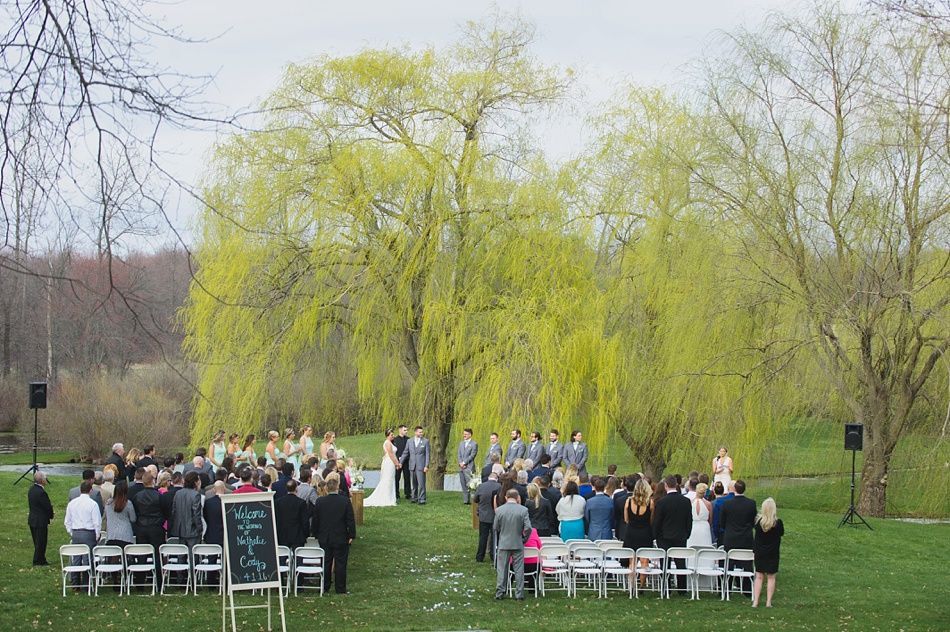 Wedding ceremony outdoors, couple at altar under a weeping willow tree. Guests seated on white chairs.
