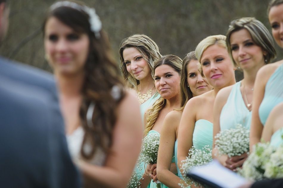 Bridesmaids in teal dresses watch wedding ceremony, some smiling and holding bouquets outdoors.