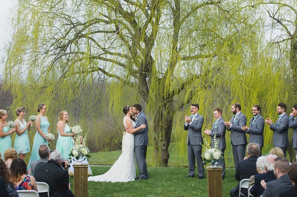 Wedding ceremony outdoors; bride and groom kissing, bridal party, guests seated, under a willow tree.
