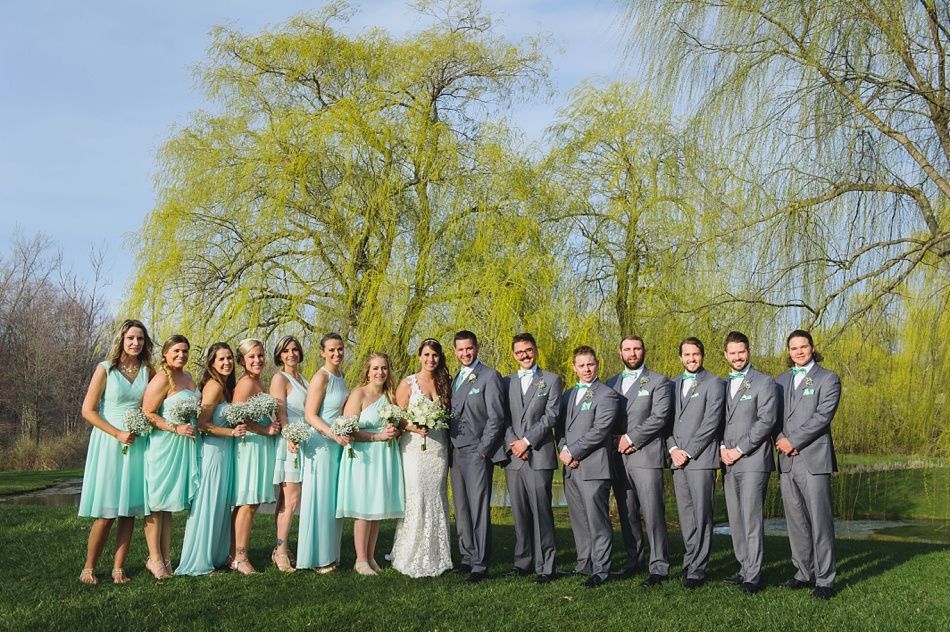 Wedding party poses outdoors in front of a tree with green leaves; bridesmaids in teal, groomsmen in gray.