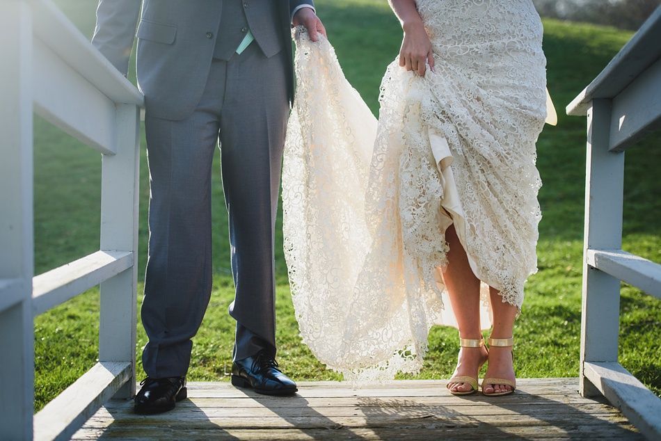Groom holding up bride's lace wedding dress train on a wooden bridge, sunny outdoor setting.