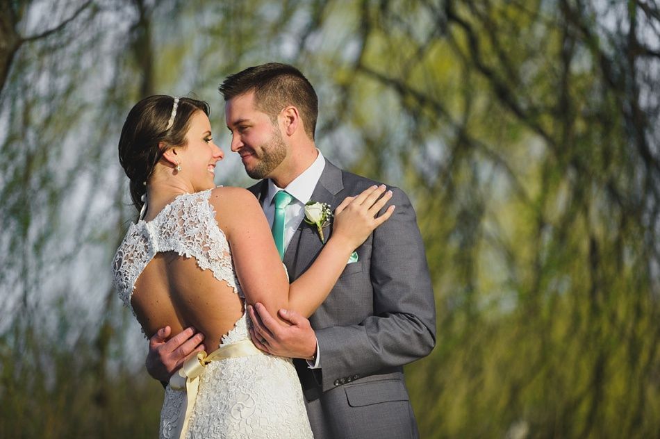 Bride and groom embrace outdoors; she wears a lace dress, he wears a gray suit and holds her.