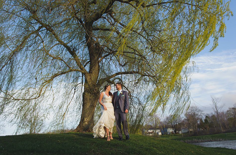 Bride and groom stand under a willow tree. Bride in white dress, groom in grey suit, smiling. Blue sky.