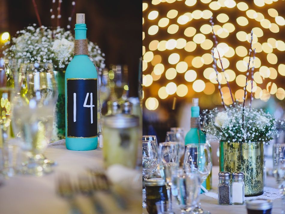 Two wedding table centerpieces: painted turquoise bottles with number 14, and gold vase with white flowers.