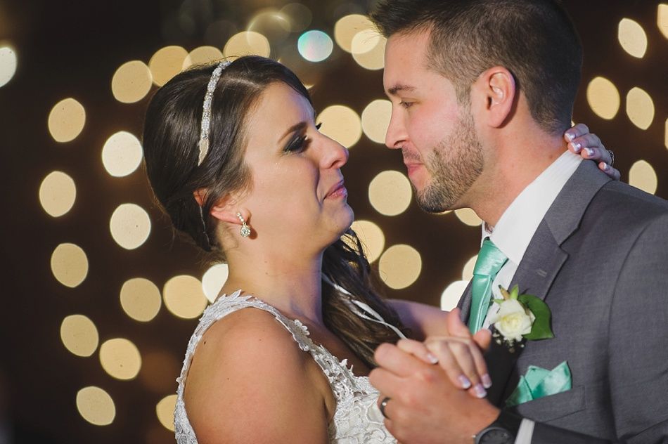 Wedding couple dancing, bride emotional, groom smiles. Bright lights in background.