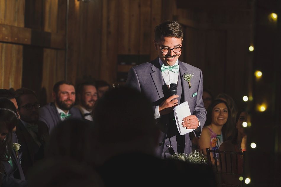 Man in gray suit speaks at a wedding reception; others seated, smiling. Wooden interior, string lights.