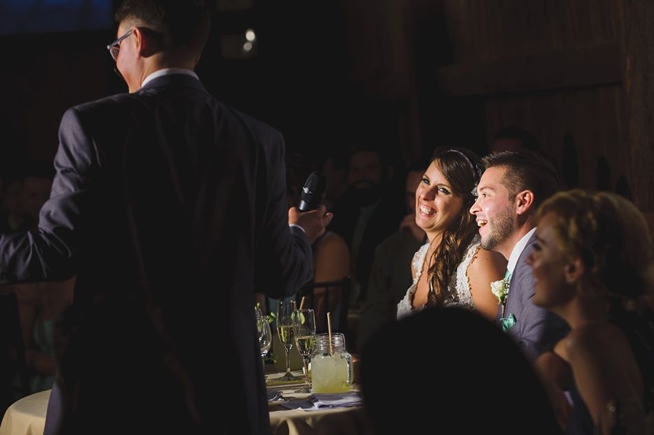 Man giving a speech at a wedding, bride and groom seated, looking on.