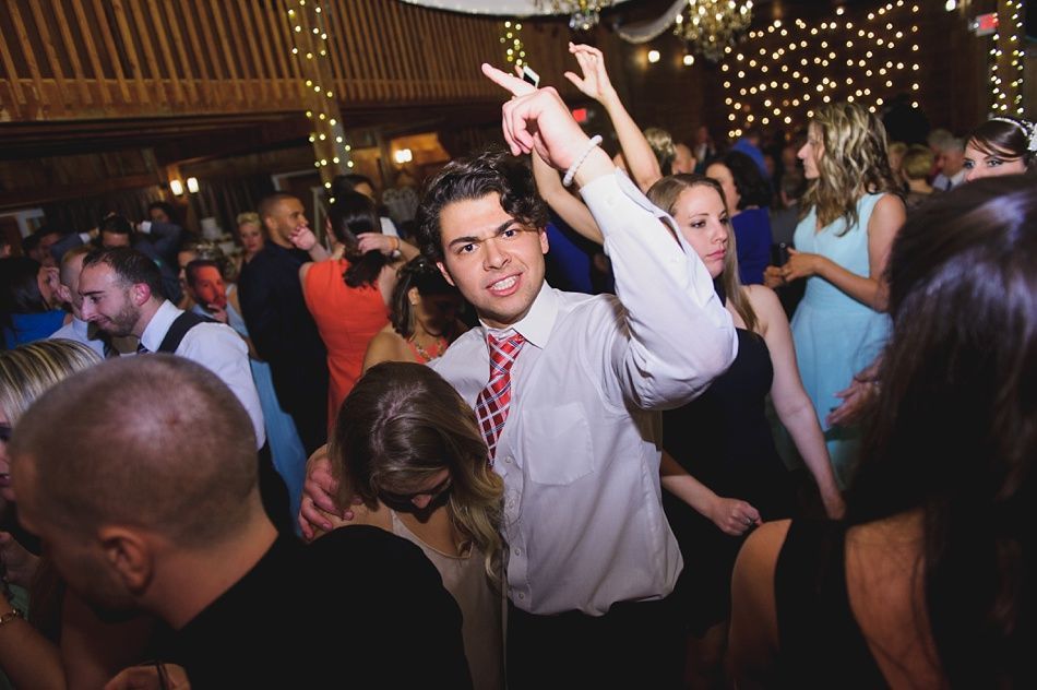 Man in white shirt dancing at a party, arms raised, pointing. Other people in the background, dimly lit.