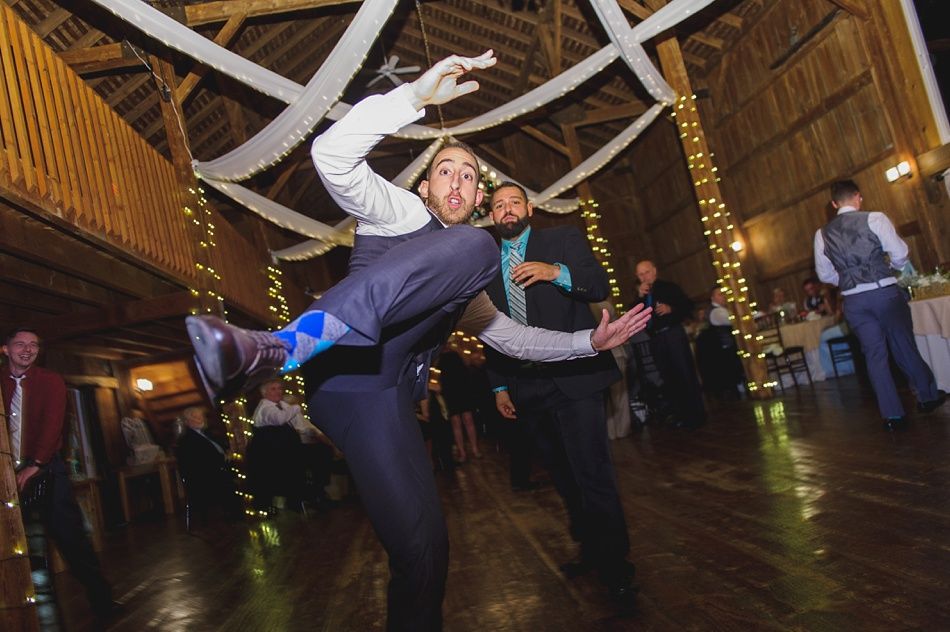 Man in suit mid-air, joyful expression, dancing in a barn-like reception venue. Blue socks visible.