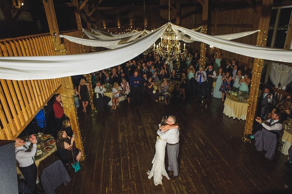 Bride and groom dance at reception in barn with guests watching. White fabric drapes from the ceiling, wood floor.