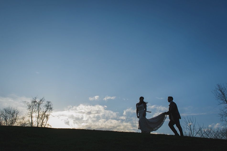 Silhouette of a couple holding wedding dress train on a hilltop against a blue sky.