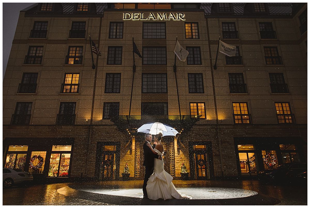 Bride and groom under umbrella in front of Delamain hotel at night, illuminated by rain and store lights.