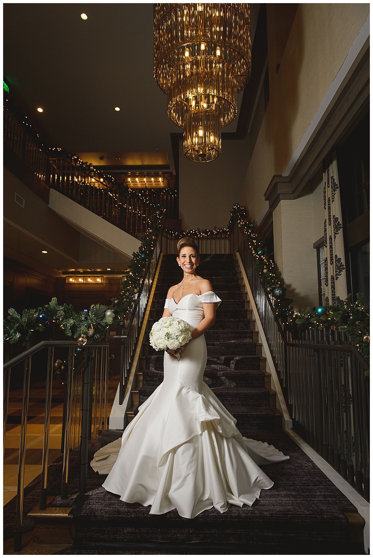 Bride in a white gown holding a bouquet on a grand staircase, under a chandelier.