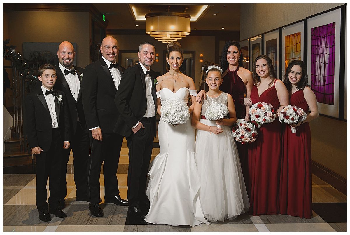 Wedding party posing indoors: bride in white dress, bridesmaids in red, groomsmen in black tuxedos.