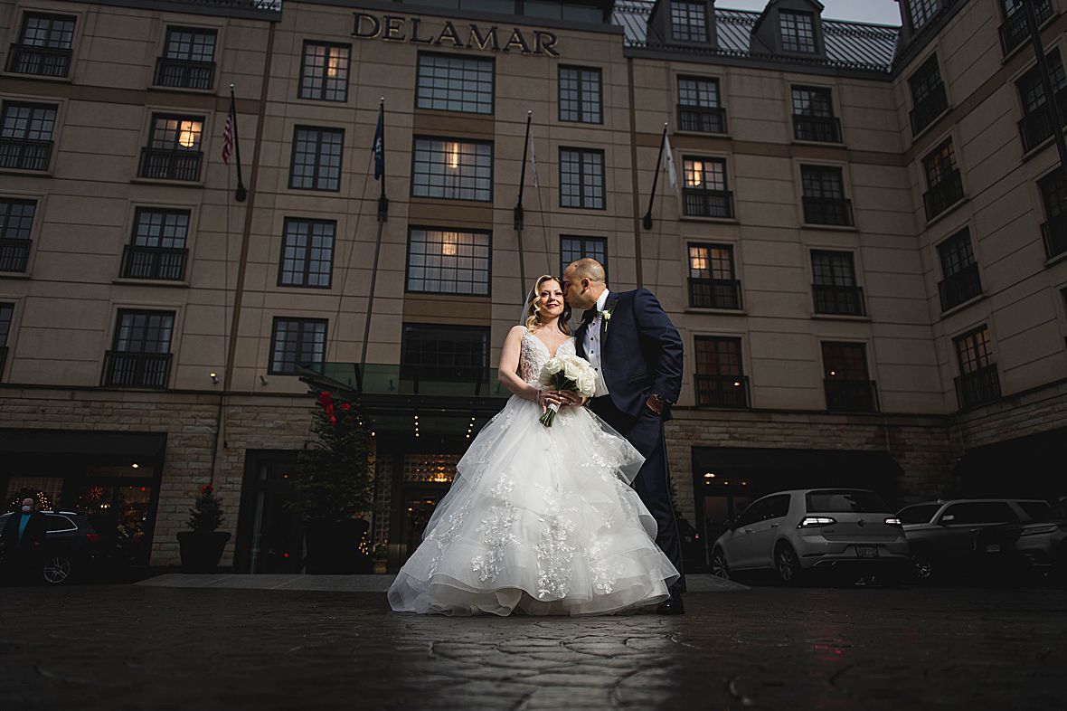 Bride and groom pose in front of the Delamar hotel. She wears a white gown, he wears a suit. Rain-slicked pavement.
