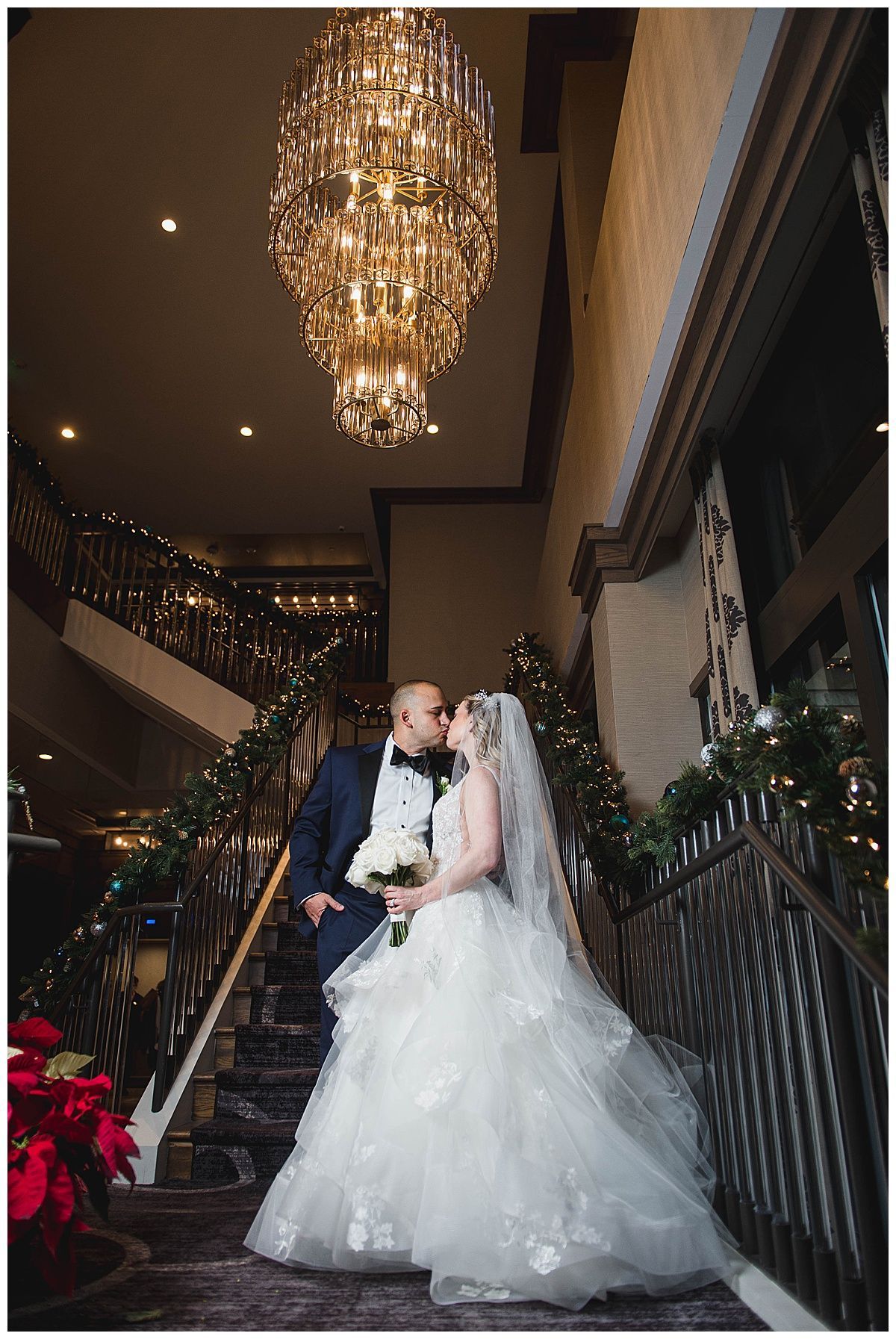 Bride and groom kissing on a staircase, ornate chandelier overhead. Festive greenery, dark suit, white gown.