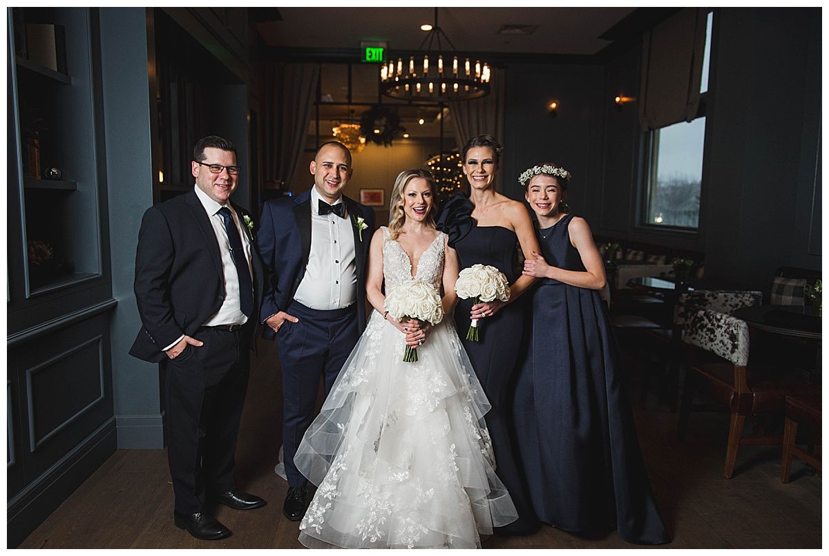 Wedding party posing indoors: bride in white dress, groom in suit, bridesmaids in dark dresses.
