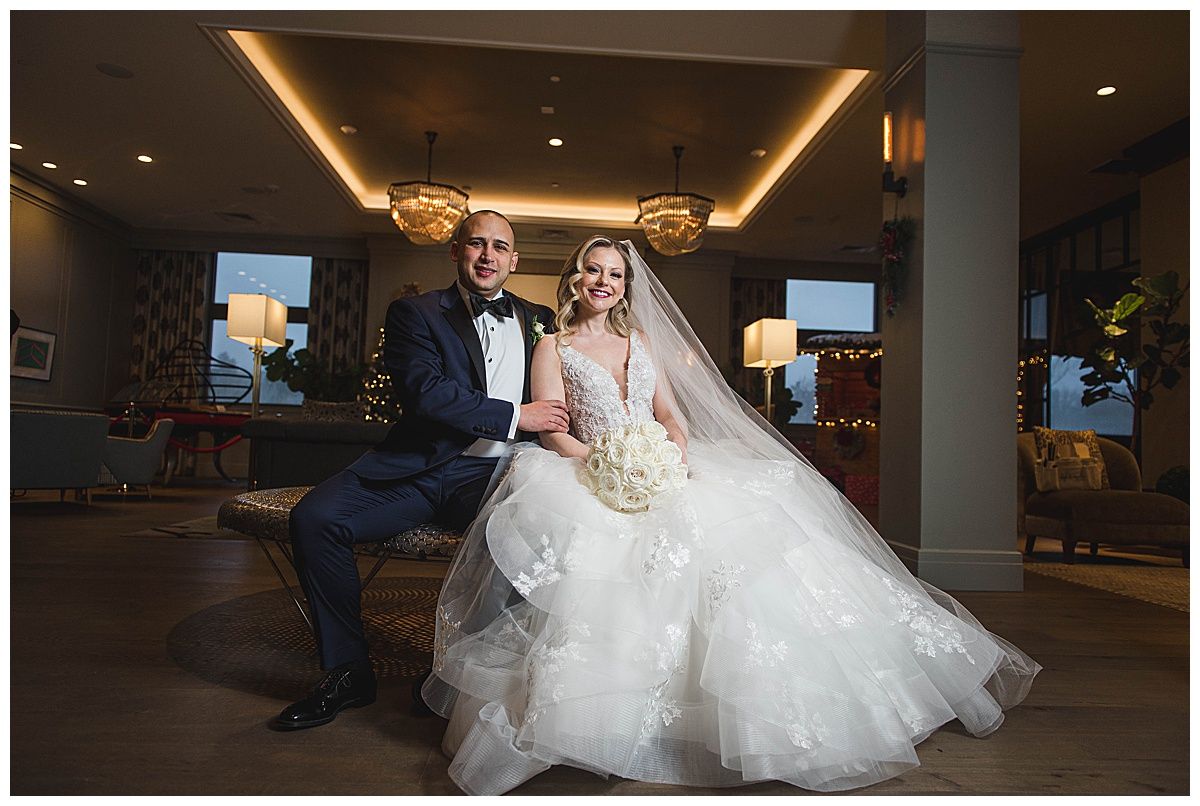 Bride in white gown and groom in navy suit pose indoors. She holds a bouquet, he sits on a bench, smiling.