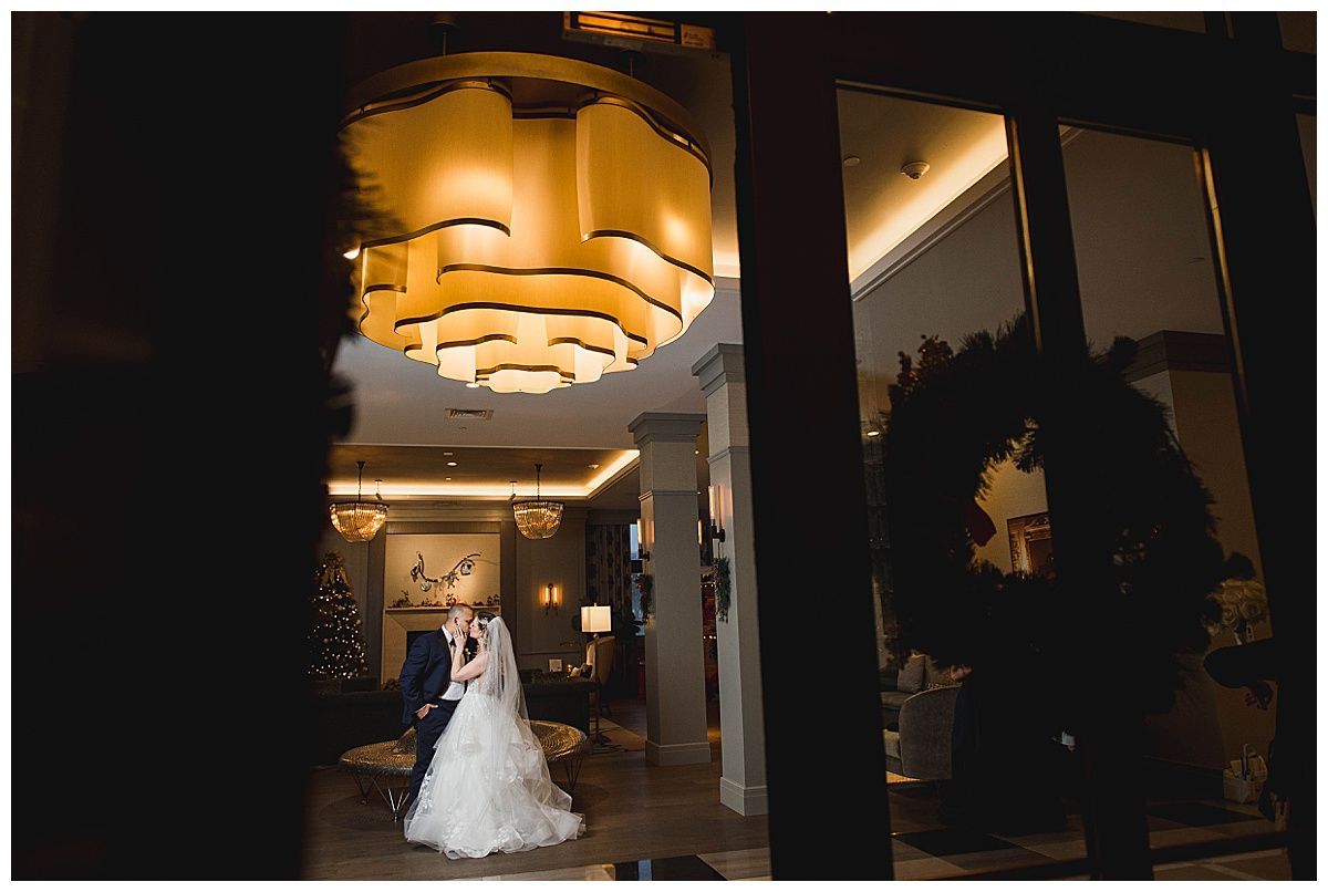 Bride and groom embrace in a brightly lit hotel lobby, viewed through glass doors with a wreath.